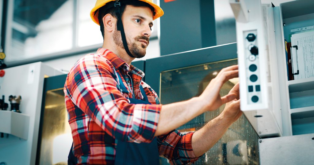 Factory worker with a yellow safety hat on punching buttons on a machine while standing up.