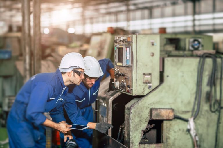 two workers looking into a machine one with a tablet both wearing blue uniforms with safety goggles and helmets